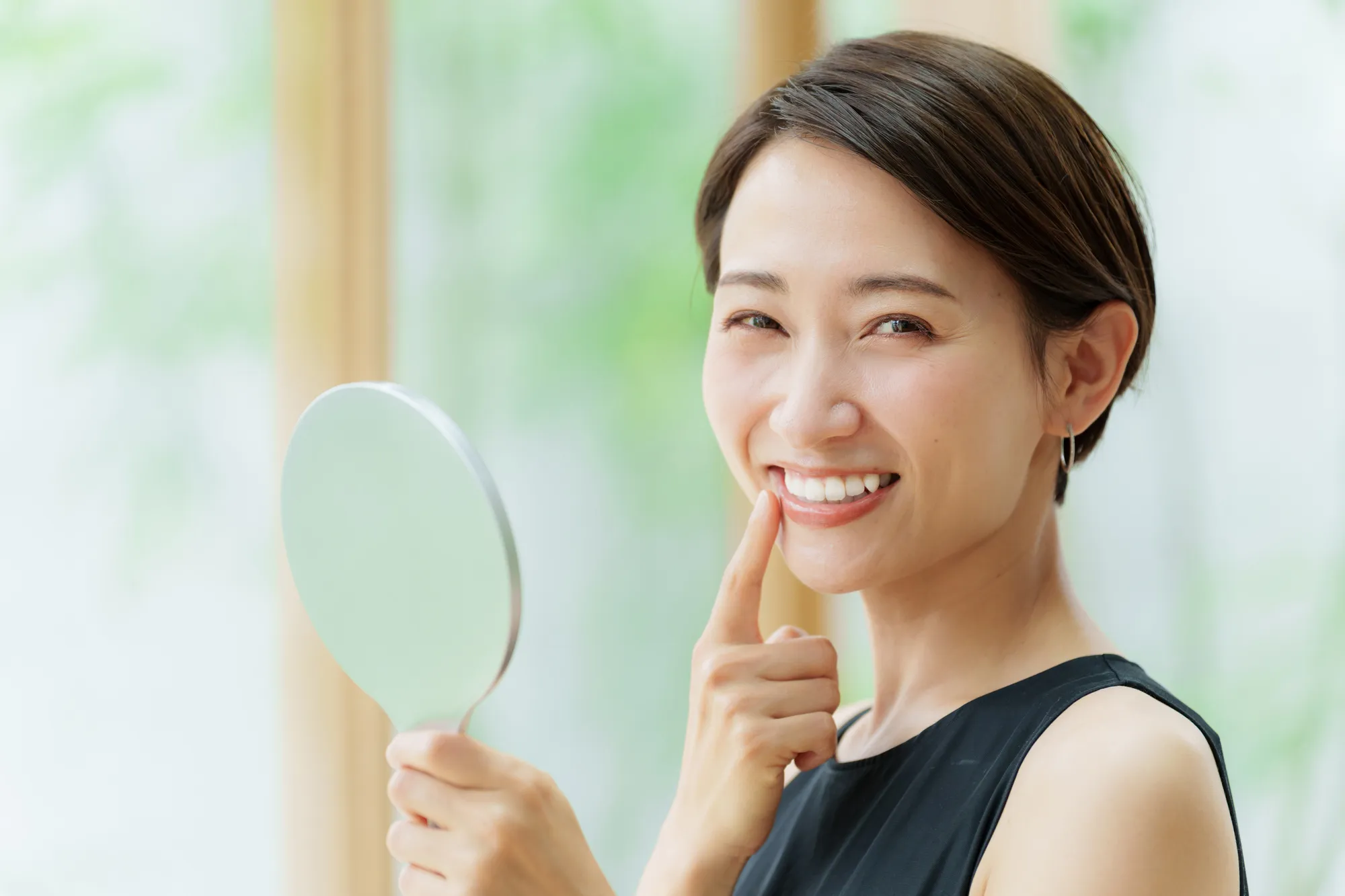 Smiling woman in black sleeveless top holding a hand mirror and touching her cheek.